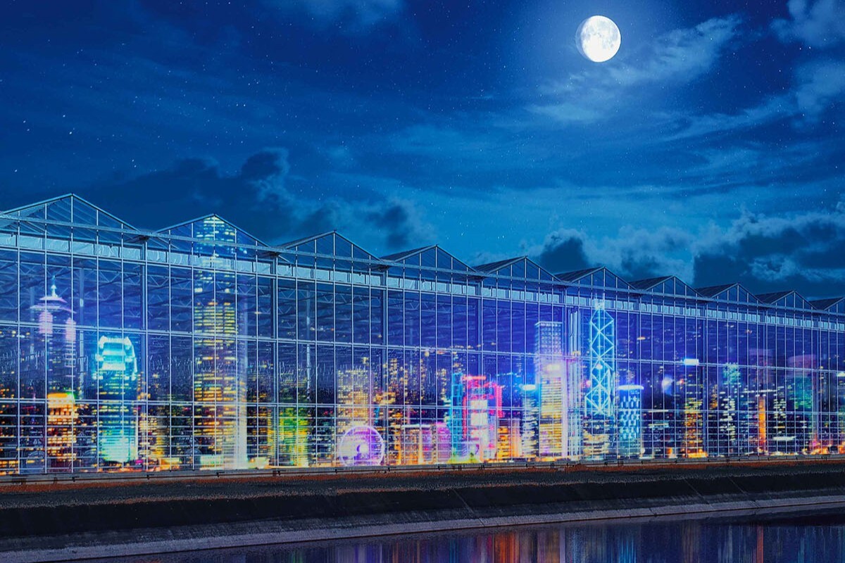 A greenhouse at night reflecting a brightly lit city skyline with tall buildings, under a dark sky with a full moon, and the city and greenhouse reflections visible in the water in front.