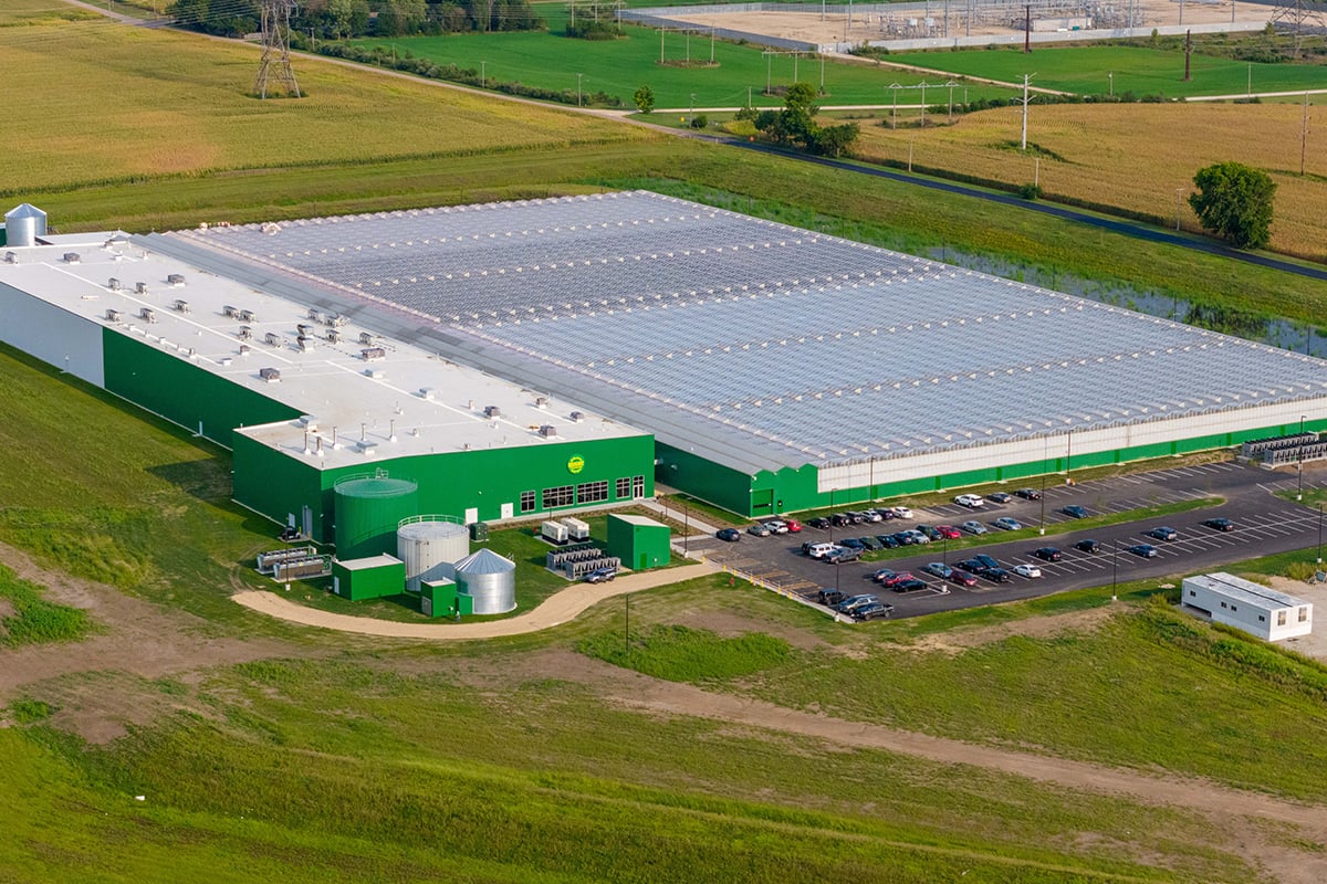 An aerial view of a large greenhouse. There are multiple white tanks, a green silo, and a green spraying system attached to the building. In front of the building, there is a parking area with several cars and a few service vehicles. The surrounding area has patches of green grass and some trees.