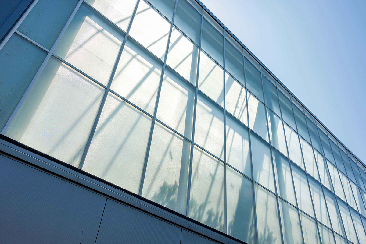 A close-up of a modern building with a facade composed of numerous large, square glass panes arranged in a grid pattern. The glass reflects the sky and some of the building's structure, creating a dynamic and contemporary look. The sky in the background is clear and blue, suggesting a sunny day.